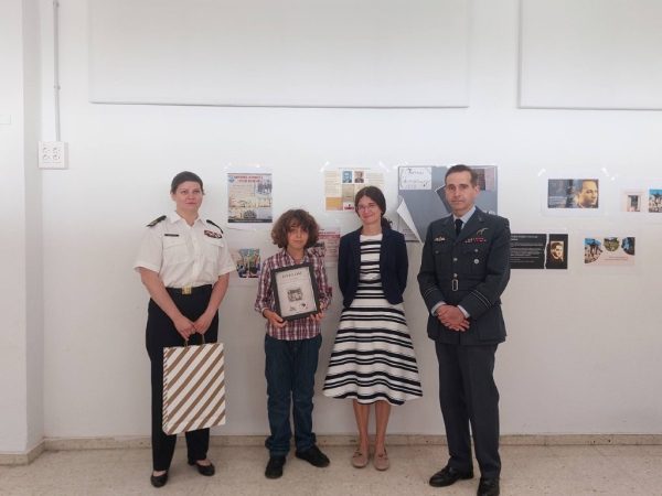 Four people stand in a hallway in front of wall display boards: a woman in a white military uniform, a boy holding a framed certificate, a woman in a striped dress and blazer, and a man in a dark military uniform.