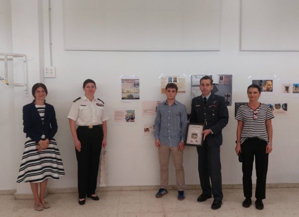 Five people stand in a gallery hallway in front of display boards; a man in a military uniform holds a framed certificate.