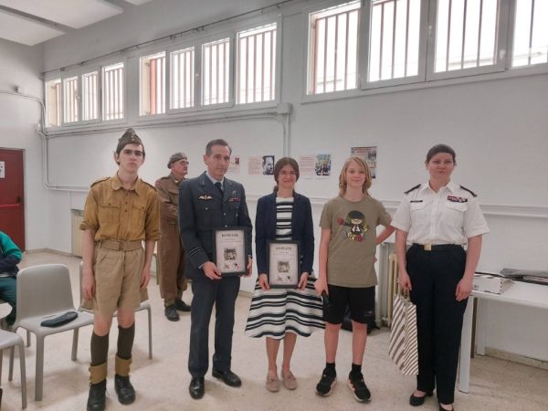 Group of five standing in a bright room, two in military dress uniforms holding diplomas, others in smart casual and a white uniform, posing for a ceremony photo.