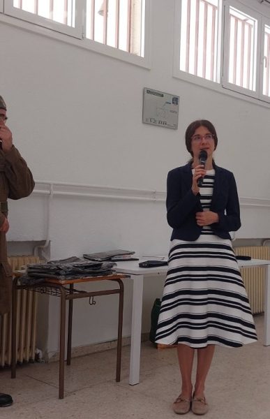 Woman in a navy blazer and striped dress speaks into a microphone in a bright room with tables and papers nearby.