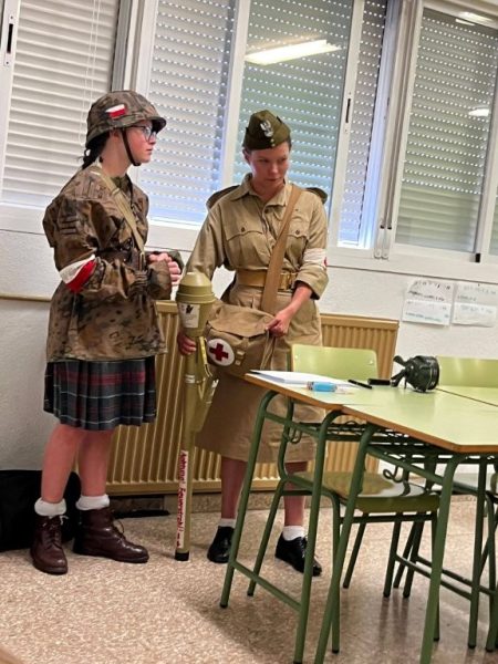Two people in vintage military costumes in a classroom; one wears a camouflage jacket and plaid skirt, the other in khaki with a Red Cross bag and a baton-like object nearby on the table