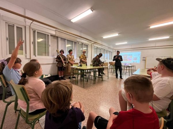 Children sit in a classroom as a presenter talks to them; several people in historical military uniforms stand by a table with equipment. A projector screen shows a slide in the background.