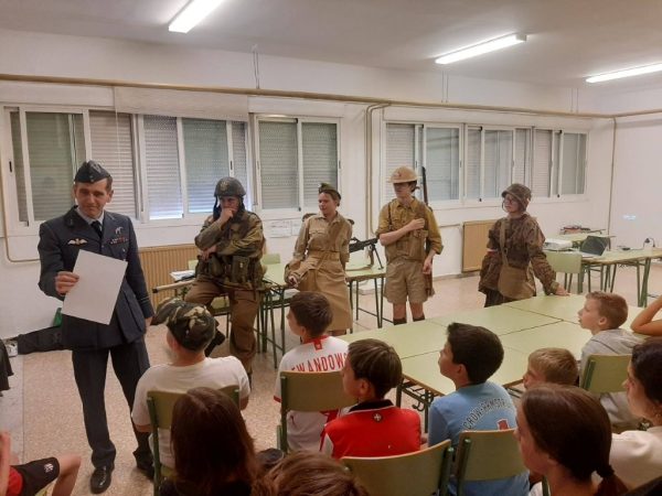 Man in a dark military uniform holding a folder speaks to children seated in a classroom; five people in WWII-era uniforms stand nearby.