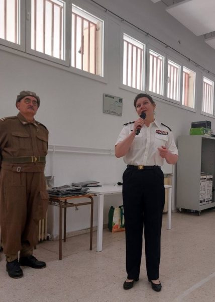 Female officer in a white uniform speaks into a microphone beside a man in a brown vintage military outfit in a bright room with large windows.