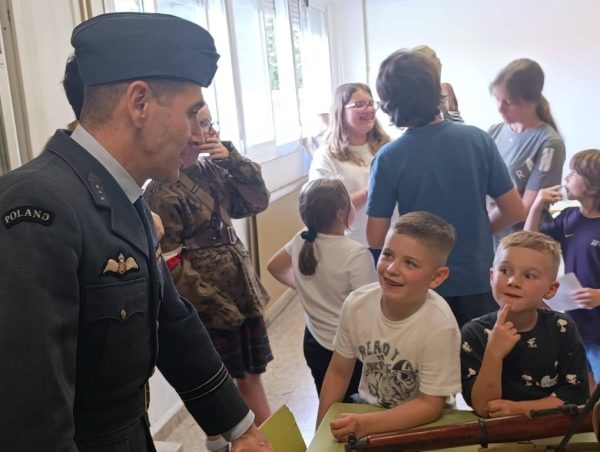 Polish soldier in dress uniform greets smiling schoolchildren in a bright hallway; a wooden rifle rests on a table nearby.