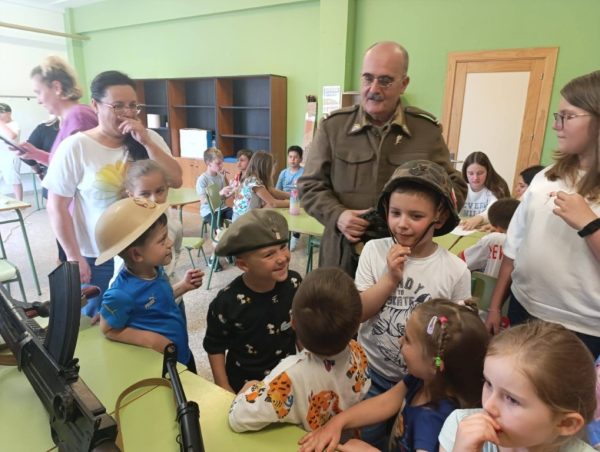 Group of smiling children wearing helmets gather around a table in a green classroom, while a man in a military-style uniform watches nearby.