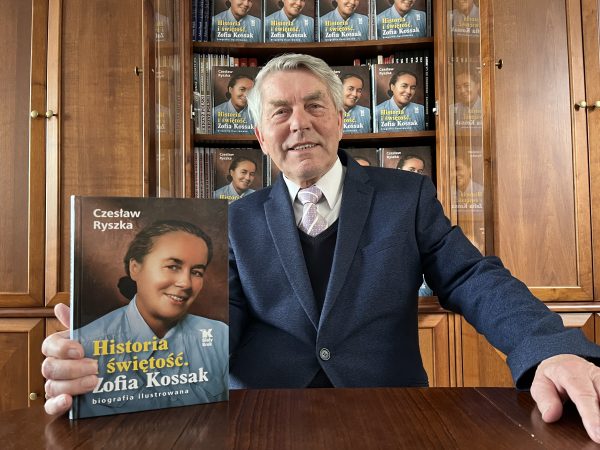 Man in a blue suit holds up a Polish biography book about Zofia Kossak in a wood-paneled library backdrop, smiling at the camera.