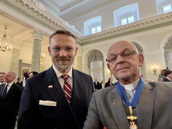 Two men in formal suits take a selfie at a ceremonial event in an ornate marble hall with chandeliers and tall columns.