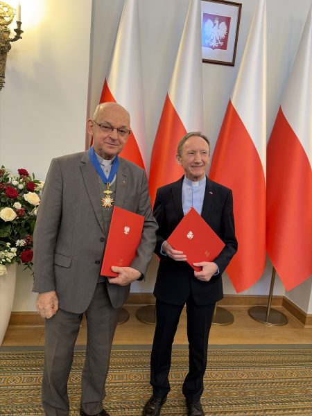 Two men in suits posing indoors with Polish flags; one wears a medal and blue ribbon, holding a red folder..