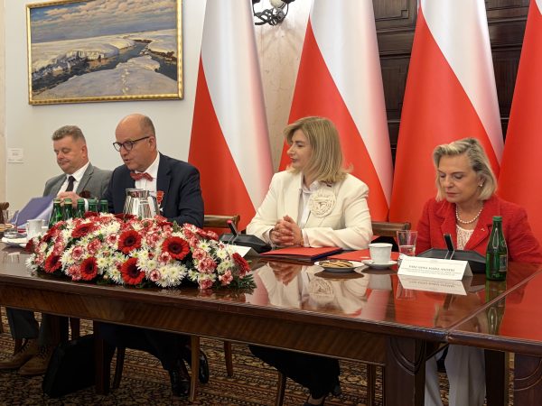 Three officials sit at a formal meeting table, with red-and-white flags behind them and a large red-white flower centerpiece in the foreground.