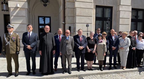 Group of formally dressed officials and military personnel standing in a row outside a stone building during a formal ceremony.