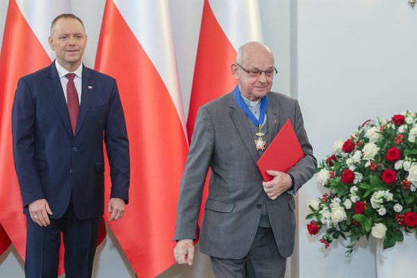 Two men in formal suits at a ceremony with red and white flags behind them; one holds a red folder beside a flower arrangement.