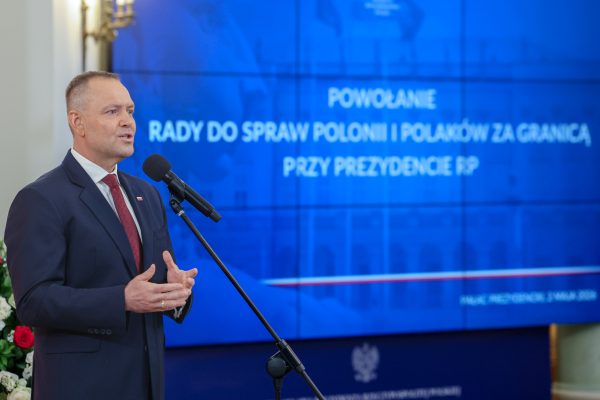Man in a navy suit speaks at a podium, microphone in front, against a blue backdrop with white text.