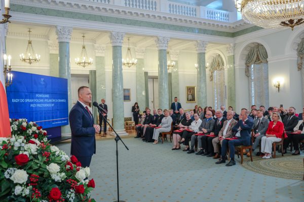 Politician in a dark suit speaks at a podium in a grand marble hall with a blue banner and formally dressed attendees watching.
