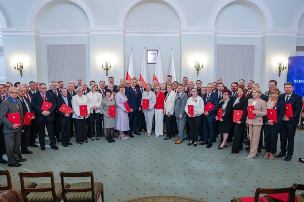 Group of formally dressed people posing with red folders at a ceremonial event in a grand room with Polish flags in the background