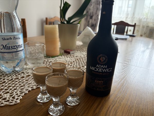 Six crystal shot glasses arranged on a wooden table around a dark Adam Mickiewicz liqueur bottle; water bottle and plant visible in the background close by.