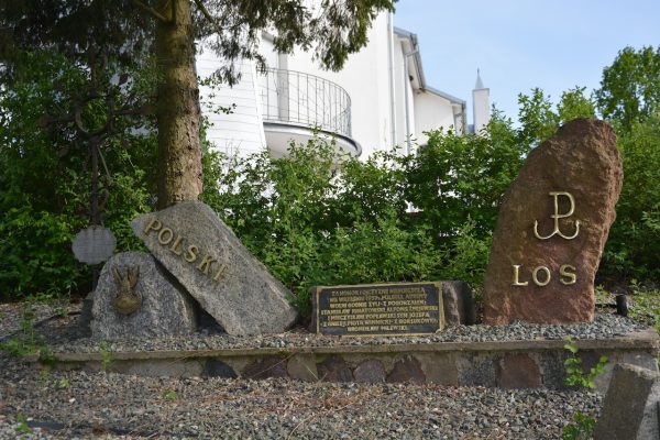 Outdoor Polish memorial with three stone markers, plaque in front, and a metal emblem; left stone reads 'POLSKI' and right stone bears large engraved letters, surrounded by greenery and a white building in the background.