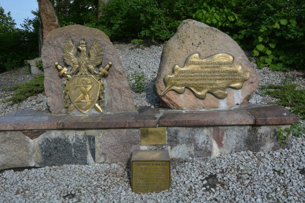 Stone memorial with two large granite blocks bearing brass plaques: left block features an eagle over a shield with swords, right block has a wavy-edged brass plaque with engraved text; both rest on a stone pedestal on gravel with green shrubs in the background.