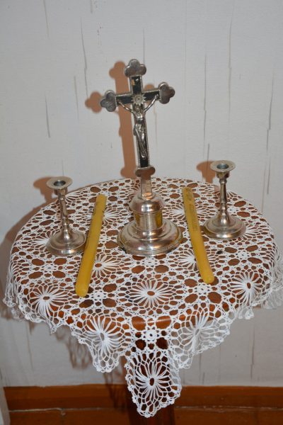 Metal crucifix on a lace-covered small table with two silver candle holders and yellow candles.