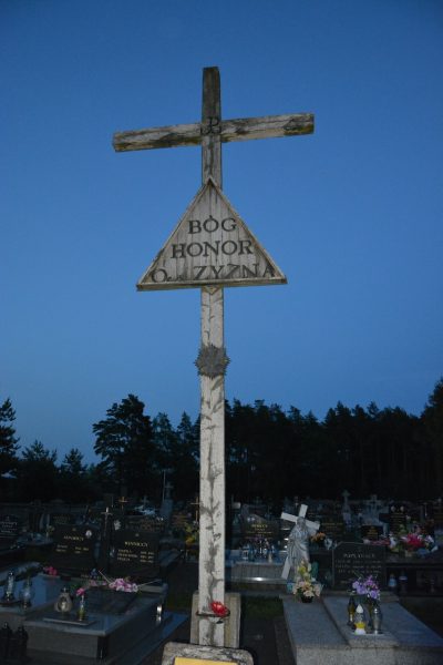Tall weathered wooden cross with a triangular plaque reading 'BOG HONOR OJCZYZNA' in a cemetery at twilight.
