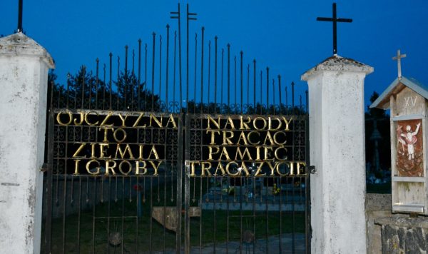 Wrought-iron gate with gold Polish inscription at a cemetery, flanked by stone columns with crosses; the text proclaims a patriotic message about homeland and memory.