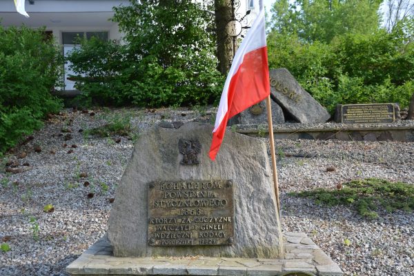 Stone memorial with a red-and-white Polish flag planted beside it in a garden setting; brass plaque honors fallen soldiers from 1863 and later generations.