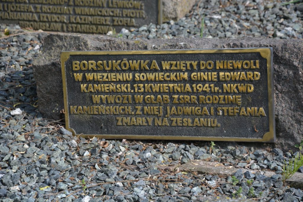 Bronze memorial plaque with Polish inscription mounted on a rough stone base, set among gravel and small rocks.