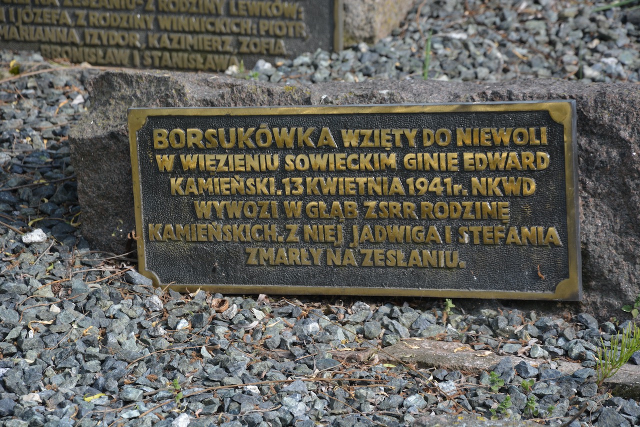 Bronze memorial plaque with Polish inscription mounted on a rough stone base, set among gravel and small rocks.