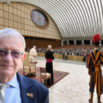 Close-up of an elderly man in a blue suit and tie with glasses, in a formal ceremony setting inside a large arched venue with a crowd and ceremonial guards in colorful uniforms.