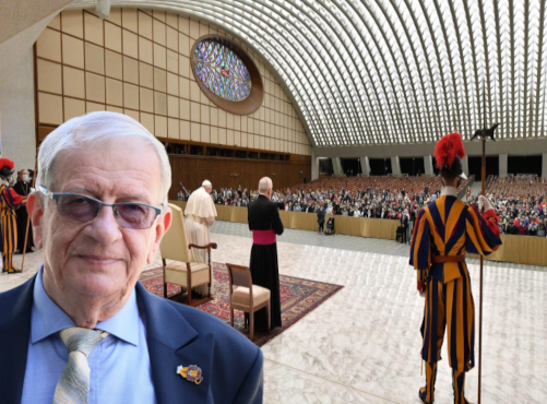 Close-up of an elderly man in a blue suit and tie with glasses, in a formal ceremony setting inside a large arched venue with a crowd and ceremonial guards in colorful uniforms.