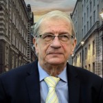 Older man with gray hair and glasses wearing a dark suit and yellow striped tie, standing on a city street at dusk.