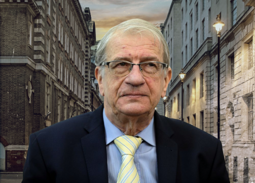 Older man with gray hair and glasses wearing a dark suit and yellow striped tie, standing on a city street at dusk.