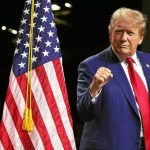 Man in a dark blue suit and red tie raising a clenched fist beside American flags on stage. Option 1: A man in a blue suit and red tie salutes with a raised fist near U.S. flags, on a stage.
