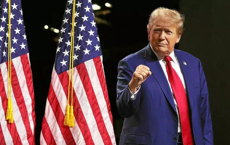Man in a dark blue suit and red tie raising a clenched fist beside American flags on stage. Option 1: A man in a blue suit and red tie salutes with a raised fist near U.S. flags, on a stage.