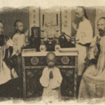 Young child stands in prayer before an ornate altar, surrounded by seated and standing adults in traditional robes or garments.