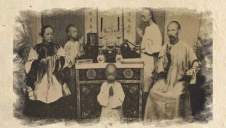 Young child stands in prayer before an ornate altar, surrounded by seated and standing adults in traditional robes or garments.