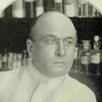 Black-and-white portrait of a man in a white lab coat with shelves of bottles in the background