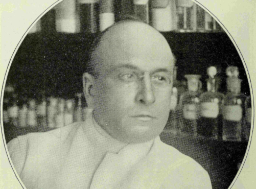 Black-and-white portrait of a man in a white lab coat with shelves of bottles in the background