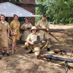 Six people in khaki World War II–era uniforms pose with vintage rifles under a white canopy.