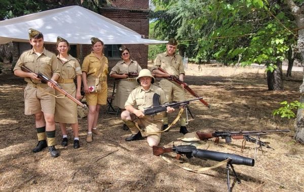 Six people in khaki World War II–era uniforms pose with vintage rifles under a white canopy.