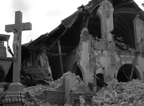 Crucifix stands amid rubble and the ruins of a collapsed church, with shattered brick arches and debris scattered around.