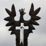 Ornamental metal sculpture of two birds with numerous signposts bearing Polish village names against a cloudy sky.