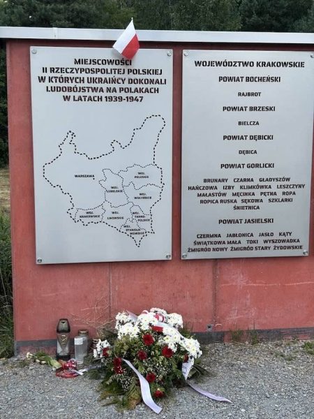 Memorial plaque panels with a map of Poland and regional names, a small Polish flag, and a bouquet of white and red flowers at the base.