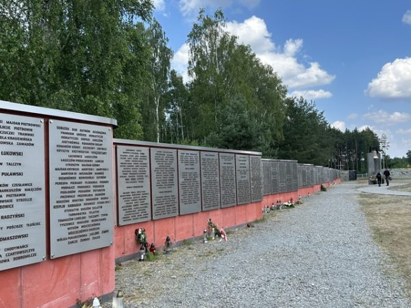 Long row of metal plaques mounted on a red wall along a gravel path, listing names at a memorial site with trees and blue sky in the background.